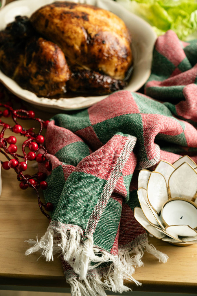 Cream holly tea towel with green/red plaid stripes and fringe beside candles and dinnerware