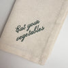 Linen napkin close-up with dark green “Eat your vegetables” embroidery and stitched hem