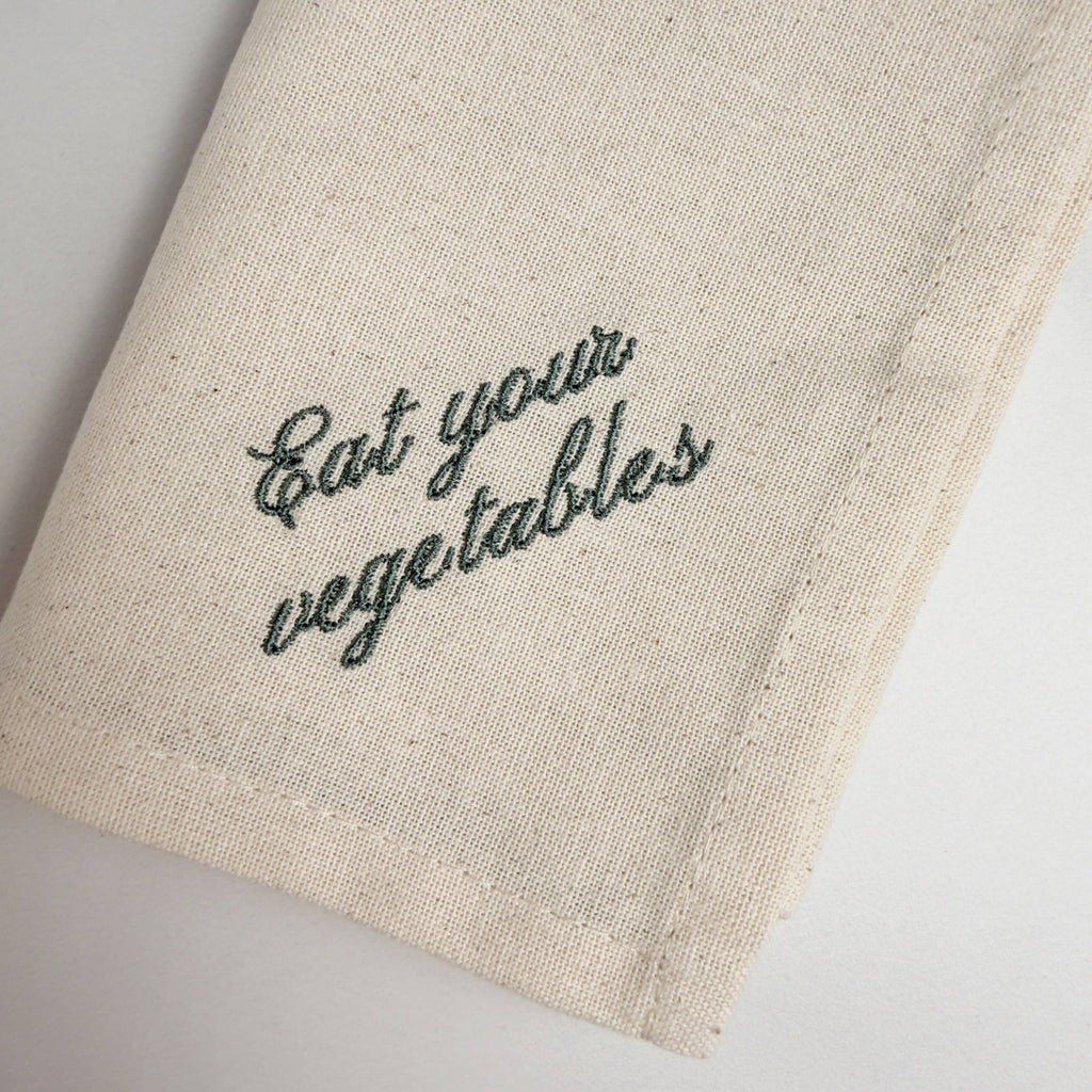 Linen napkin close-up with dark green “Eat your vegetables” embroidery and stitched hem